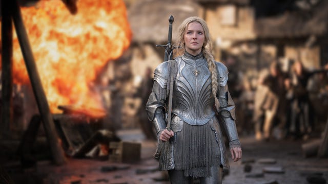 A young Galadriel standing in silver armor with a fiery explosion behind her in a village