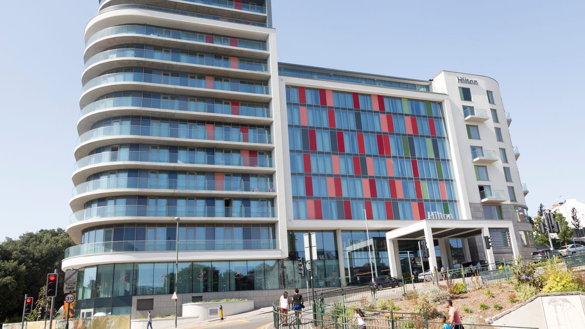 A modern Hilton hotel building with red and blue windows sits atop a small hill