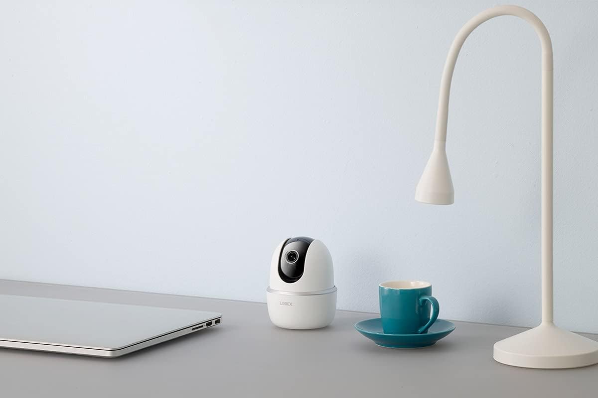 A Lorex pan/tilt indoor camera sits on a gray desk next to a closed laptop, coffee mug, and lamp.
