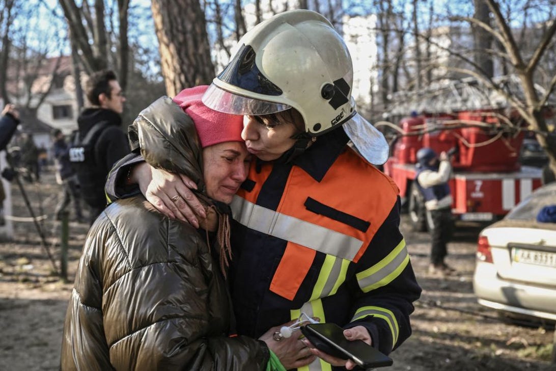 A woman in an emergency worker's uniform and helmet hugs another woman, in civilian clothes, who's crying.