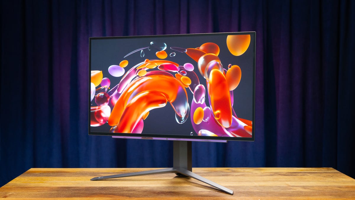 The LG Ultragear OLED 27-inch monitor angled to your left on a wood surface with a blue and purple curtain in the background and a water drop abstract on the screen