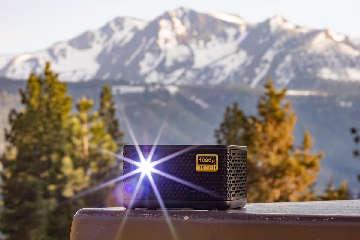 The AAXA P7+ pico projector on a table with trees and mountains in the background.