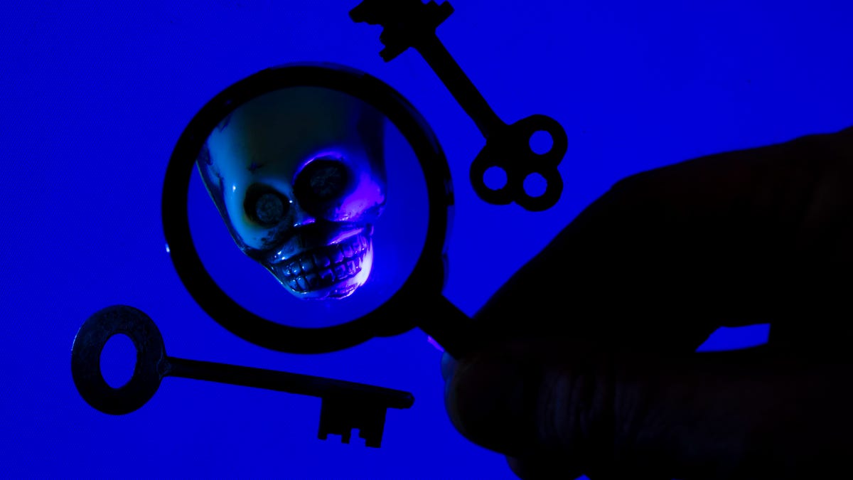 On a blue background, two old fashioned keys are displayed on either side of a shadowy hand holding magnifying glass, which reveals a magnified skull.