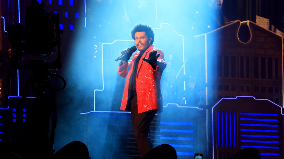 The Weeknd stands in the beam of a bright spotlight, wearing a vivid red jacket.
