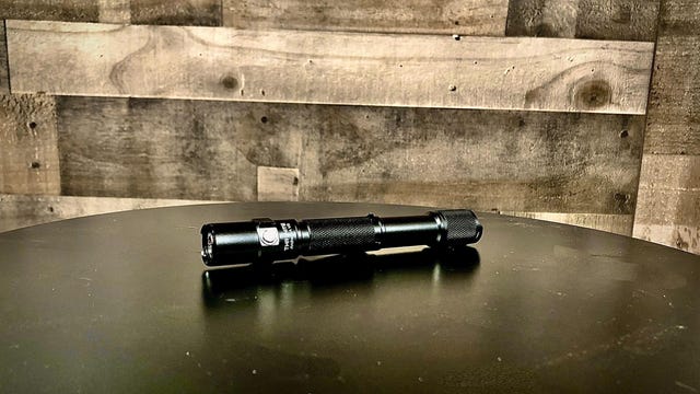 The ThruNite Archer 2A V3 flashlight sits on a black tabletop in front of a wooden wall.