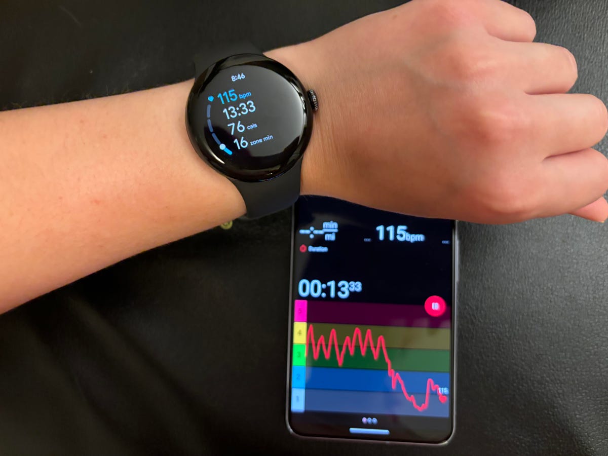A Pixel Watch 2 on someone's wrist with a Pixel phone in the background showing heart rate data
