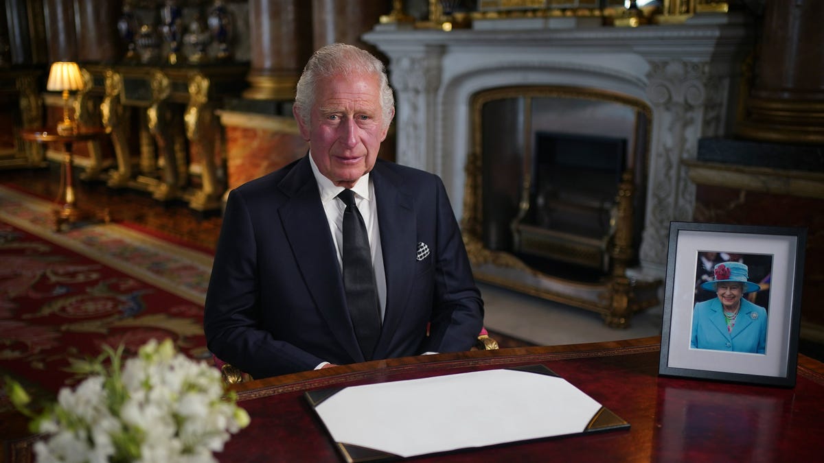King Charles III sits at a desk next to a picture of Queen Elizabeth II