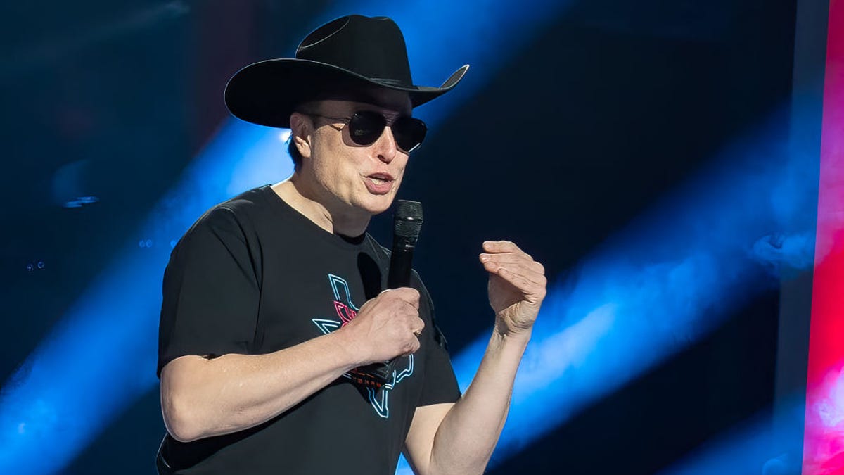 Elon Musk wears a cowboy hat and aviator sunglasses as he speaks into a microphone at a Tesla event