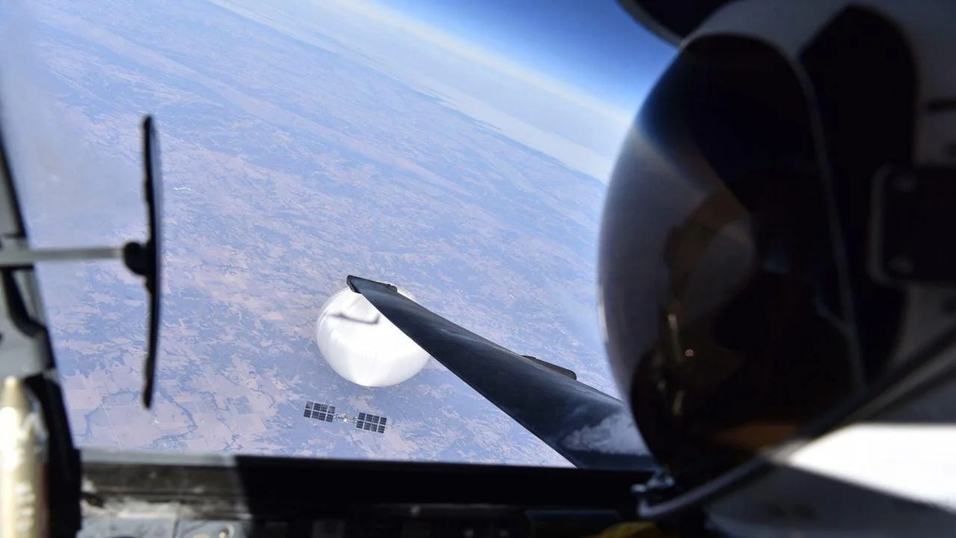 A pilot's black shiny helmet seen from the side with view forward through the window of a U-2 plane. A white round balloon floats over the US with a solar-powered payload dangling below it.