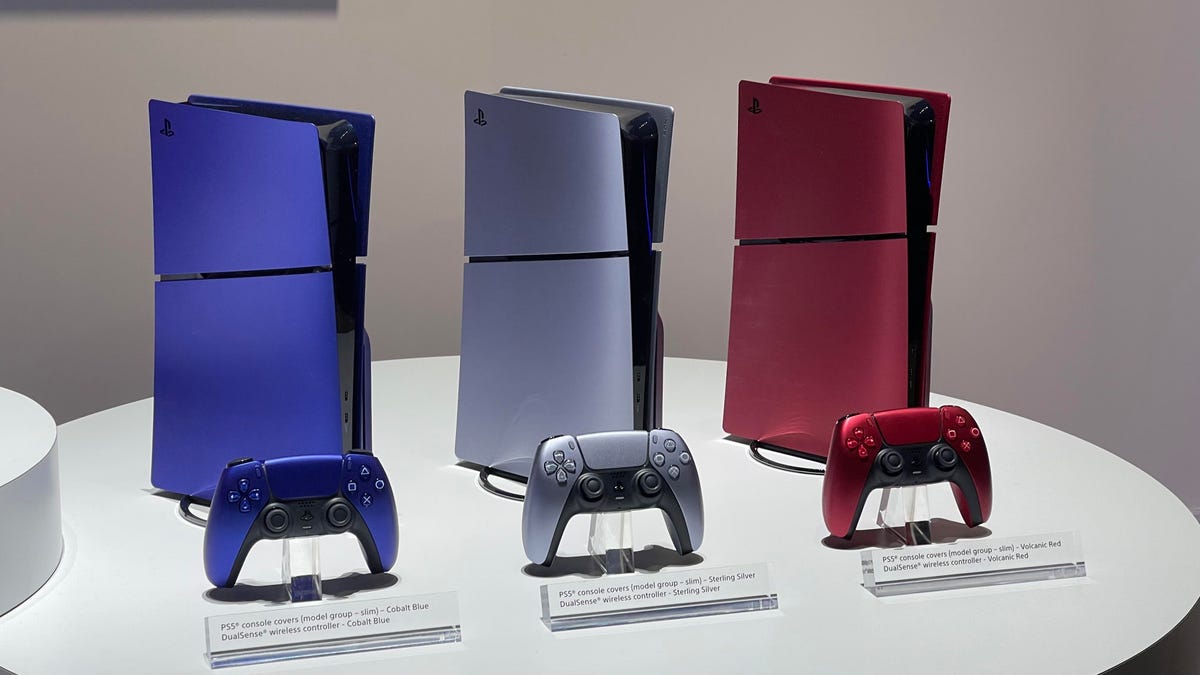Three PlayStation 5s side by side. One is blue, one is silver and the third is red. PlayStation 5 controllers are in front of each