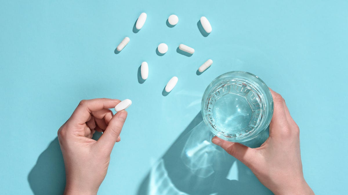 White pills on a pale blue surface, seen from above. A hand holds a single pill, and another hand holds a glass of water.