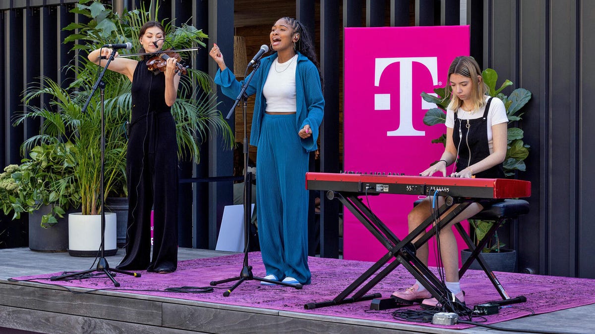 Three performers -- a violinist, a singer, and an electric pianist -- stand on a stage with a T-Mobile logo behind them.