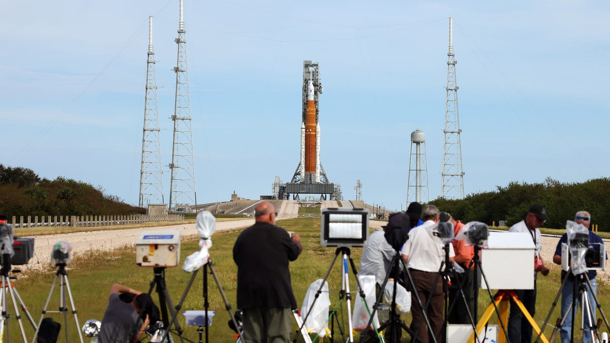 An orange-colored Artemis I moon rocket, with the white Orion spacecraft on top, is in the far ground. Skies are clear, pale blue and members of the media are setting up tripods in the foreground, facing the rocket.