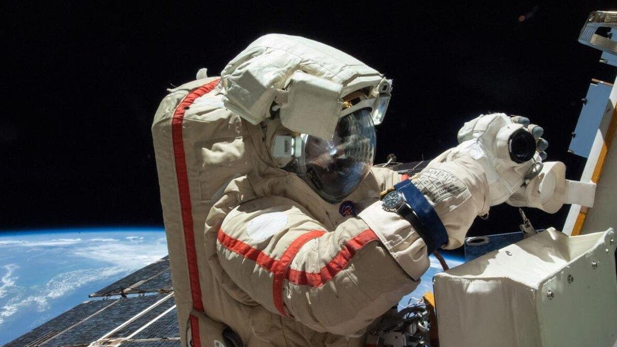 A Russian cosmonaut in a brownish-gray spacesuit with an orange stripe works outside the space station.