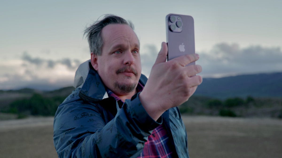 A man holding an iPhone 14 Pro outdoors on a remote hillside