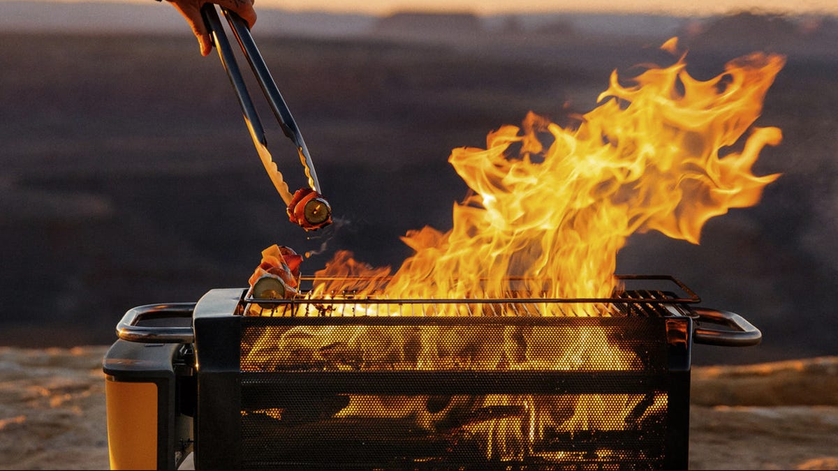 A hand with tongs reaches toward kabobs grilling on a BioLite FirePit grill.