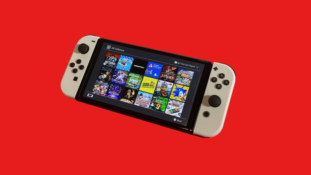 A Nintendo Switch OLED with a vast game library on the screen, against a solid red background
