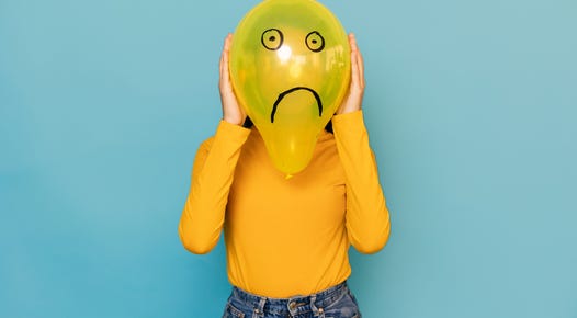 Woman hidden behing a balloon with a sad face drawn on it over blue background.