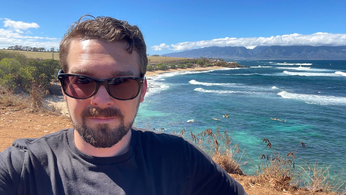 A man in sunglasses stands in front of a beach on the north side of Maui, with blue-green water lapping the golden shores.