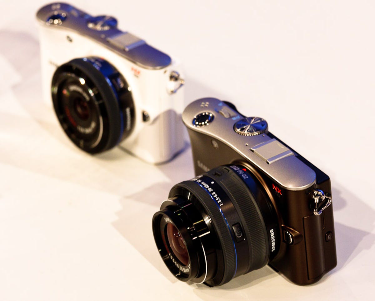 Two Samsung NX100 cameras, a white one with the 20mm f2.8 lens and a black one with the 20-50mm f3.5-5.6 lens. The camera will come in white, black, silver, and brown.