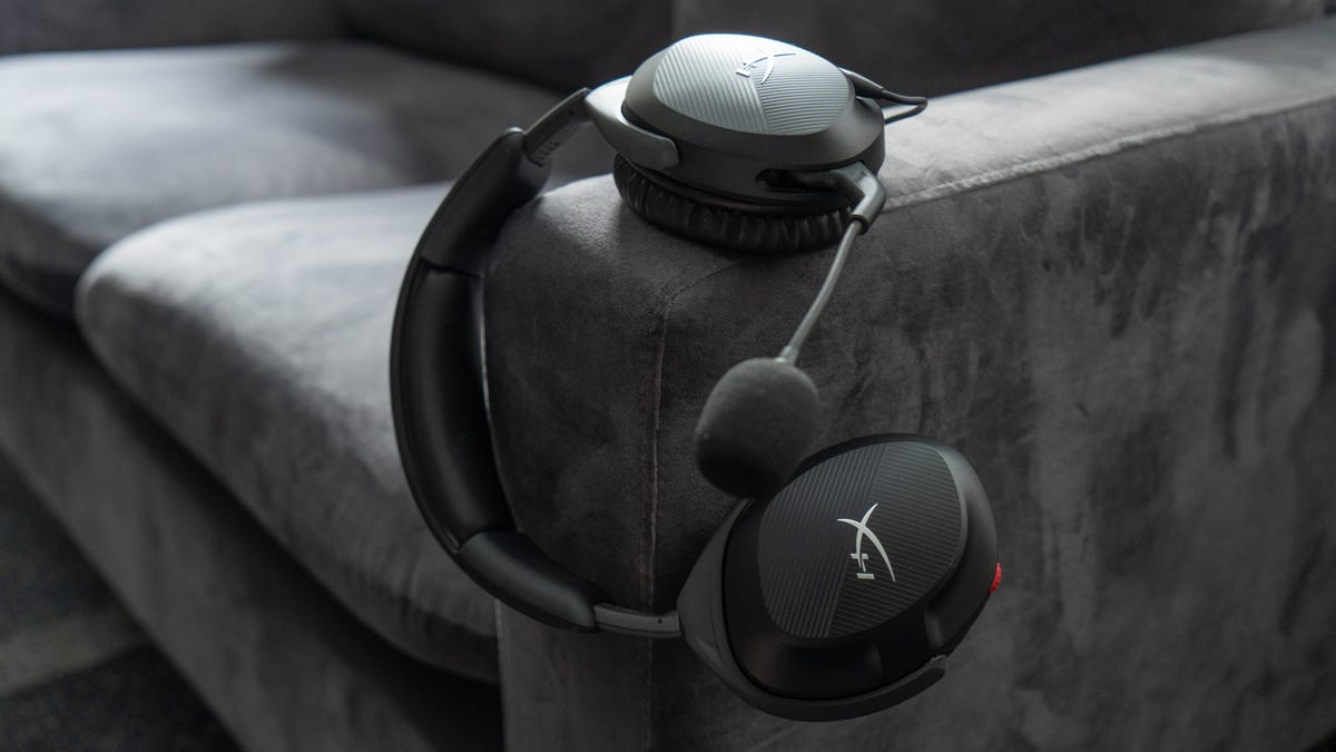 HyperX Cloud Stinger 2 hanging on the arm of a gray couch off the left earcup with the mic extended and a foam cover on the mouthpiece