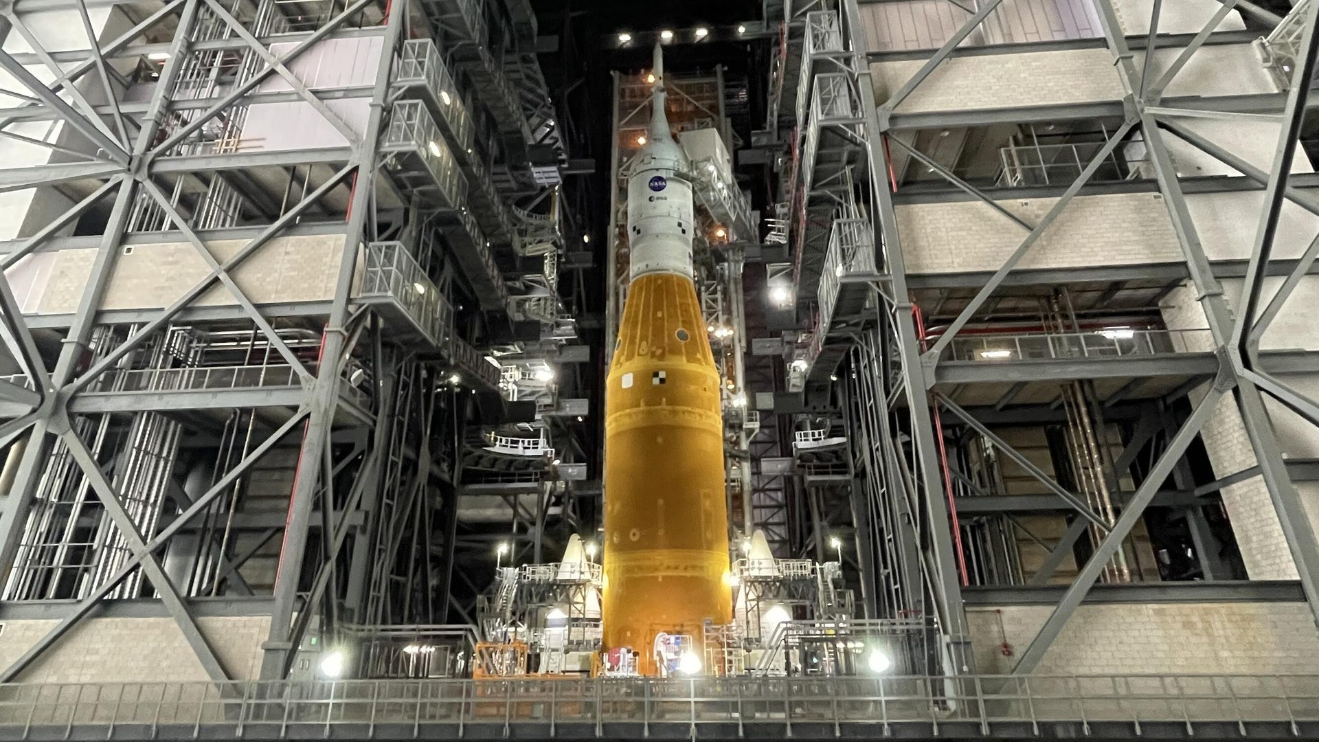 SLS and Orion, together a white capsule on top of an orange rocket surrounded by platforms inside a giant garage.