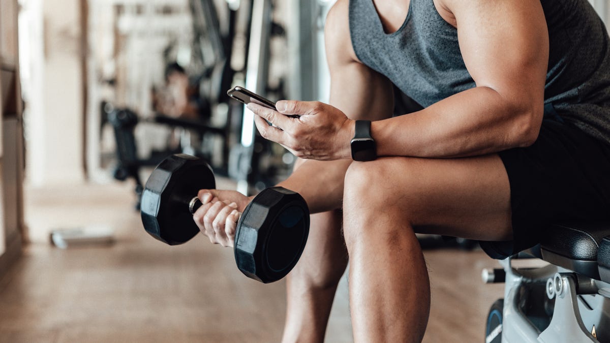 Close up shot of a young man lifting dumbbell while using smartphone