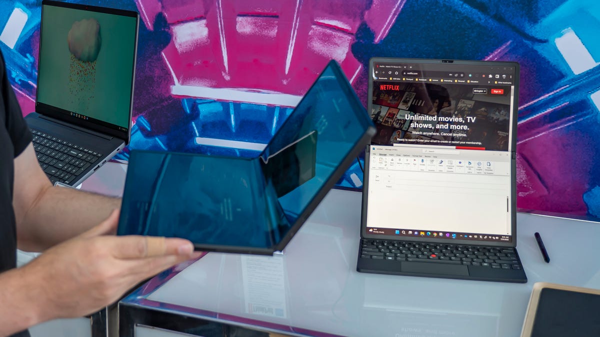 The ThinkPad X1 Fold shown in someone's hands from the side, bent in the middle to resemble a clamshell laptop, with another one to the right of it sitting on a table with the keyboard and opened to portrait orientation.