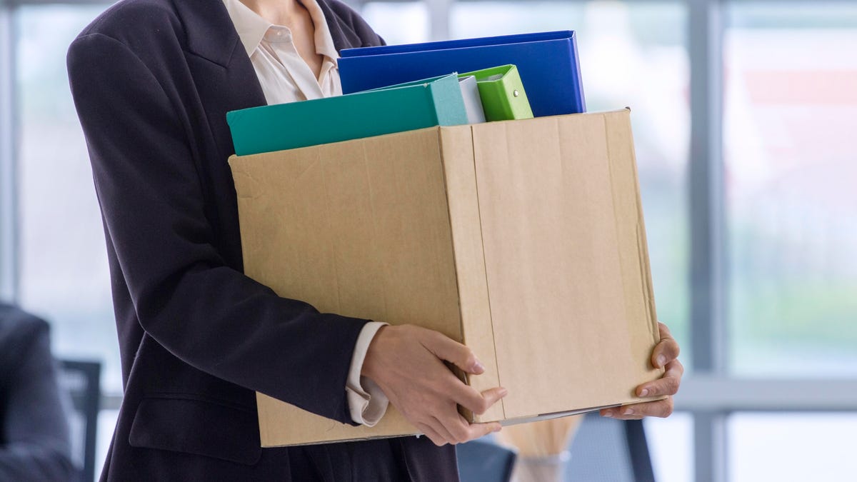 person in a suit holding a brown box with folders and leaving an office