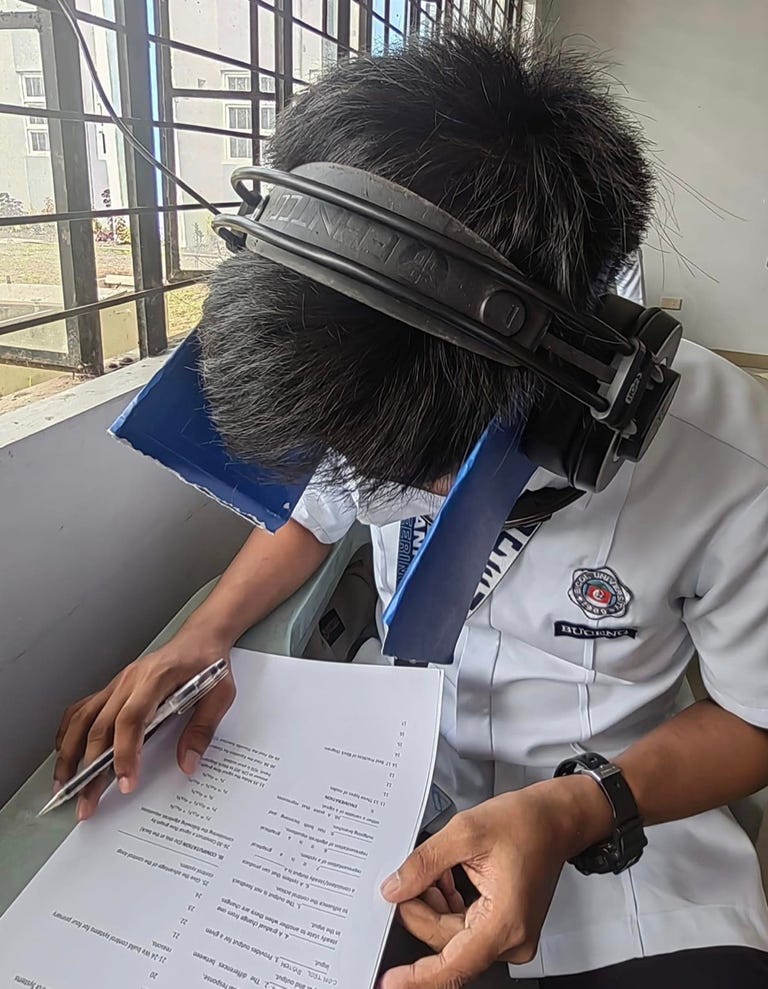 Students wears headphones to hold rectangular flaps of paper over his ears like blinders