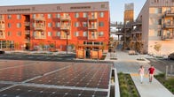 Two people walk by a large solar array in front of an orange apartment building