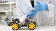 A small tabletop robot is seen with yellow wheels. A scientists is injecting a liquid into one area of the robot.