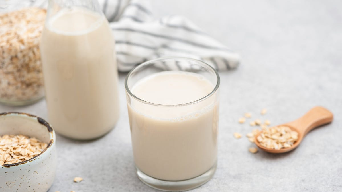 A glass of oat milk flanked by an old-fashioned glass milk bottle full of the milk and a wooden spoon overflowing with rolled oats.