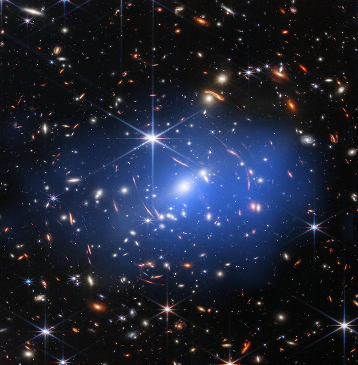Over het donkere canvas van de ruimte liggen veel kleurrijke spikkels die sterrenstelsels voorstellen.  Veel zien eruit als strepen omdat ze vanuit ons gezichtspunt vervormd zijn door zwaartekrachtlenzen.  In het midden is een blauwachtige waas die Chandra-waarnemingen voorstelt.