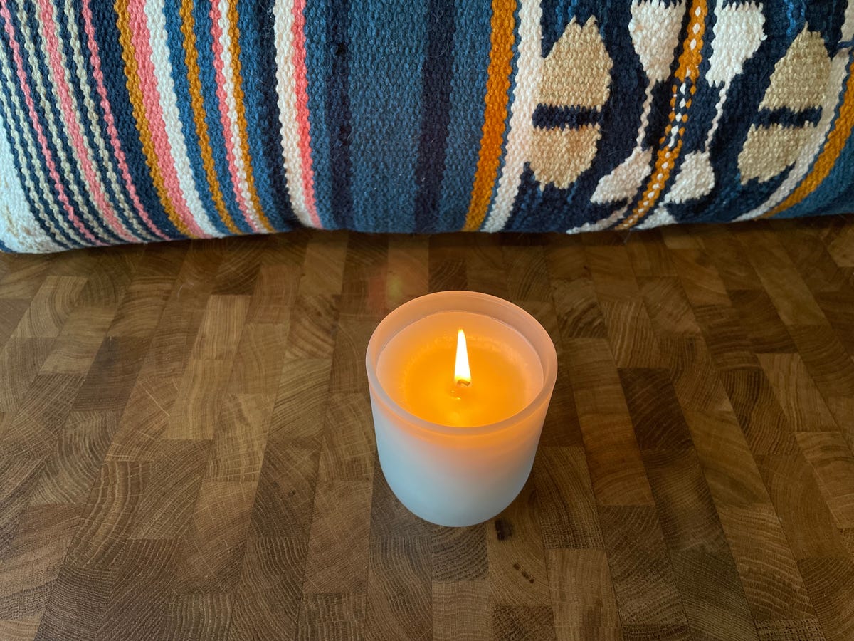 A lit candle with tunneling wax in a glass container on a wooden surface with a southwestern fabric backdrop