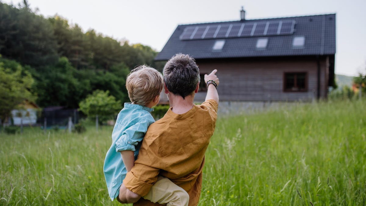 A father holds a young child while pointing at a house with solar panels on it.