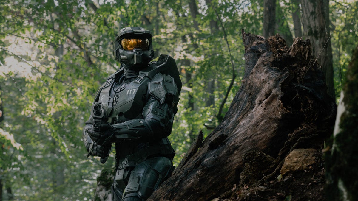 A still image from season 2 of the TV show Halo, showing the Master Chief standing next to a tree in a woodland area, holding a gun.