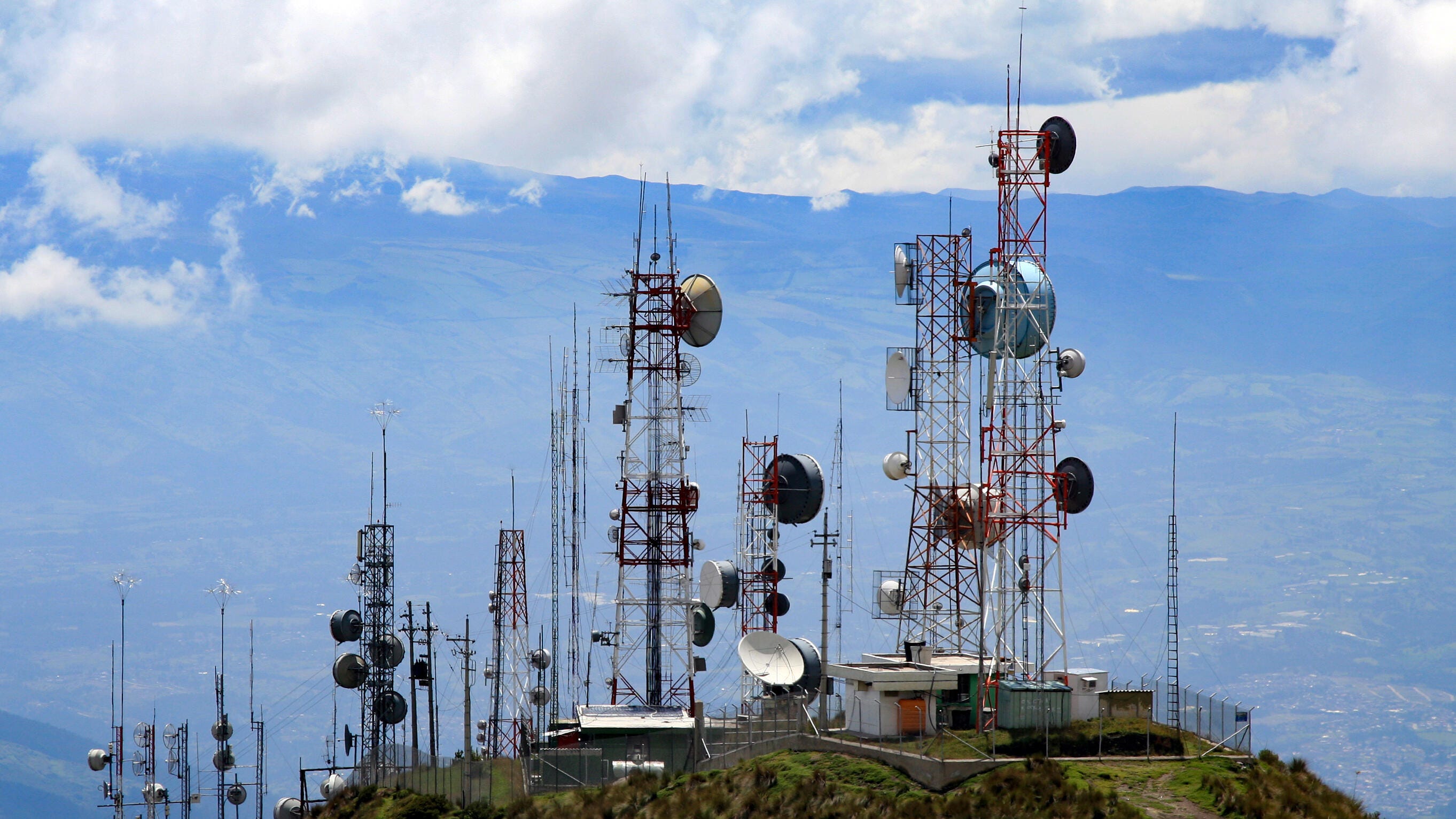 Tall towers with myriad antennae sit perched on a mountaintop.  More mountains are in the distance.