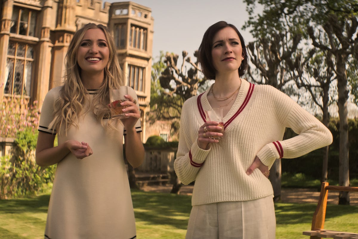 Two women standing outside a mansion wearing preppy outfits and holding drinks