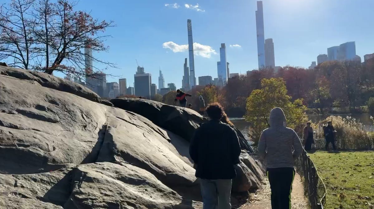 People walking around Central Park, skyscrapers in the distance, with a large rock on the left