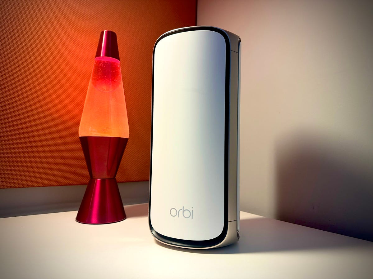 A lone Netgear Orbi 970 Series mesh extender sits on a desk in front of an orange lava lamp. It's a tall, cylindrical device roughly the size of a roll of paper towels, and it adds support for the latest-gen Wi-Fi 7 standard into the Orbi mesh mix.