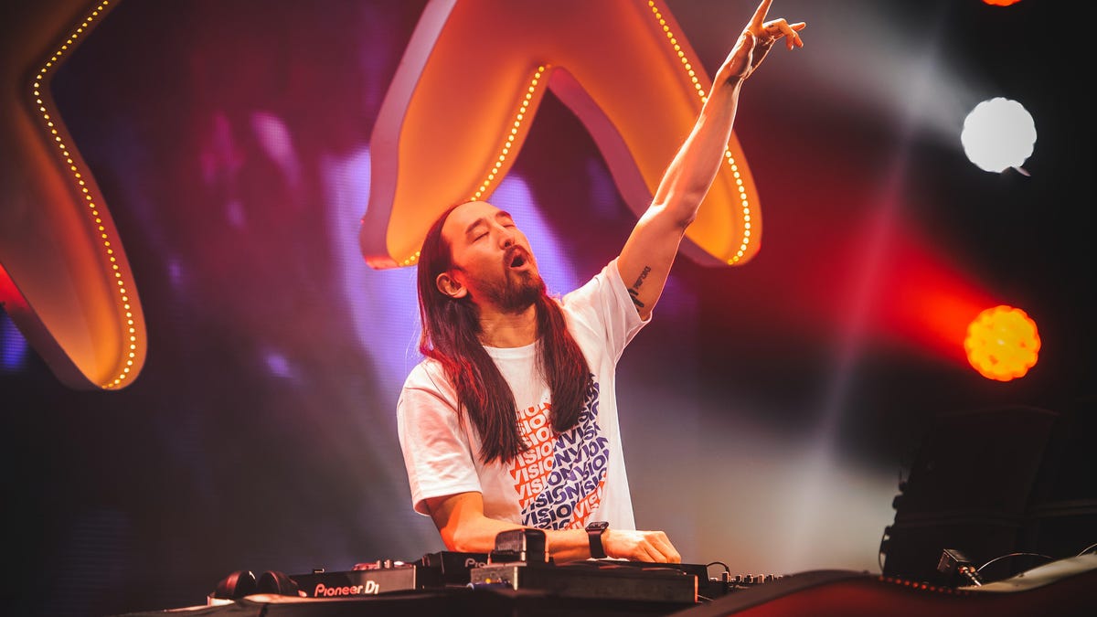 Steve Aoki lifts one hand in the air as he stands behind his mixing board