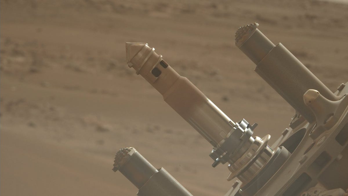 The hollow bit is tapered at the end, and features open ports on its side to allow loose material to fall inside it when the bit is inserted into Mars regolith (broken rock and dust on the surface). The blurred background shows the dusty Martian surface, with distant hills on the horizon.