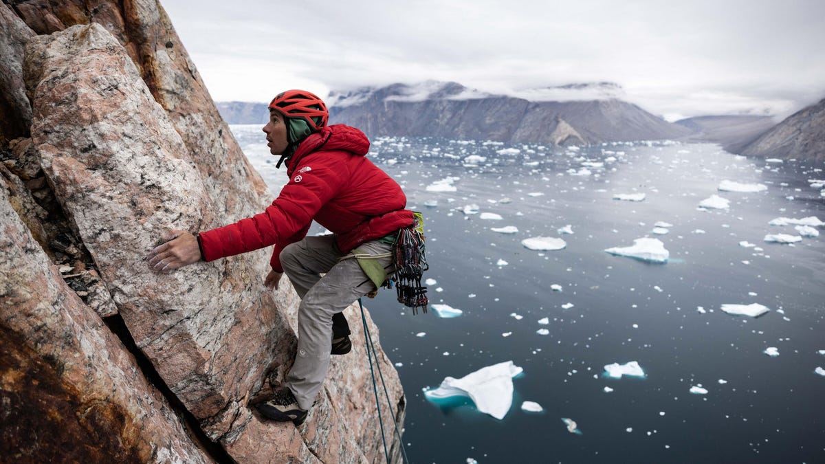A man in helmet and red jacket climbs a rock wall hundreds of feet above an ice floe-laden frigid fjord.