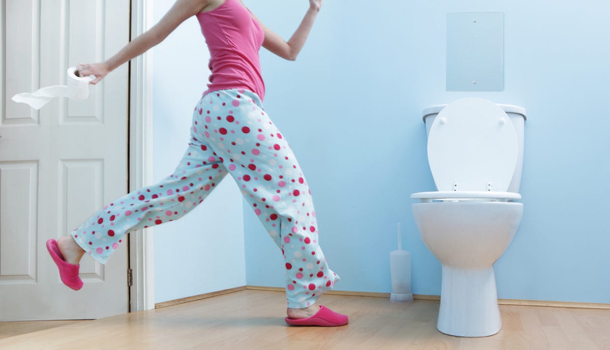 A man wearing a polka dot pajama is running towards the toilet with a roll of toilet paper in his hand.