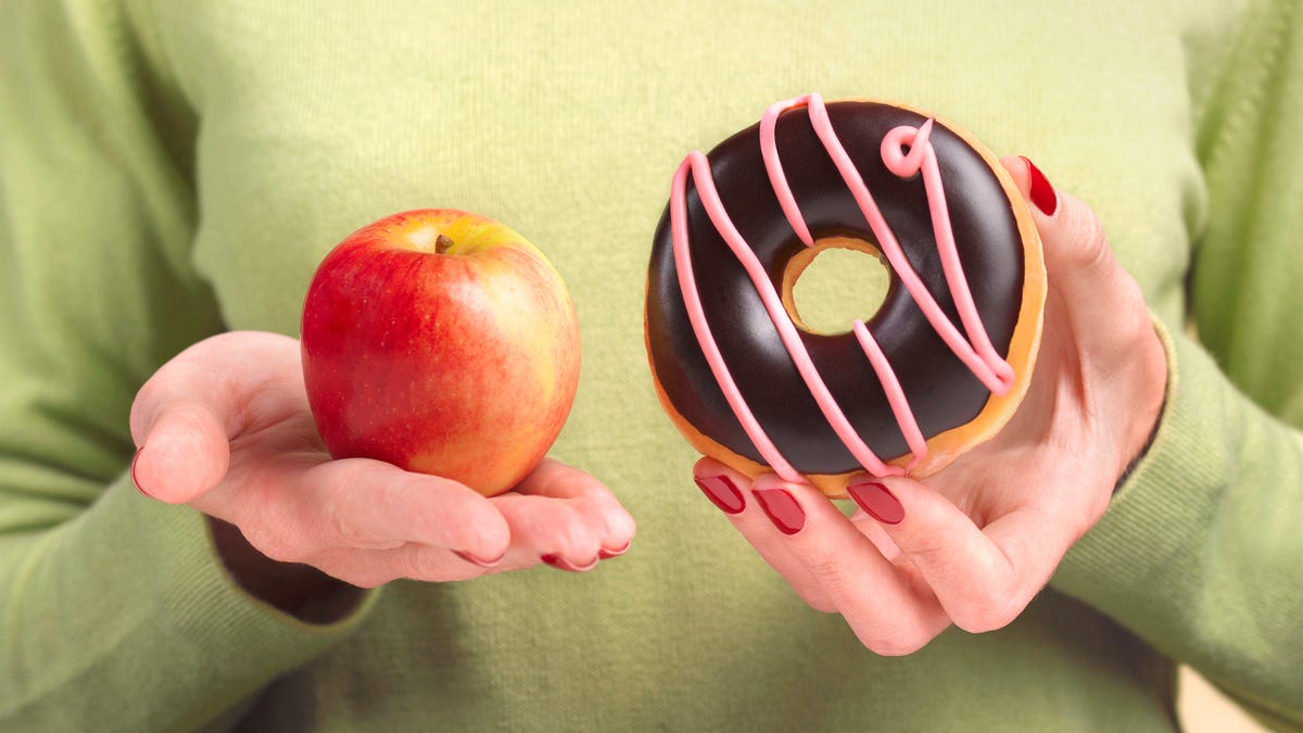 A close up of a woman's hands, one holding an apple the other hand holding a doughnut