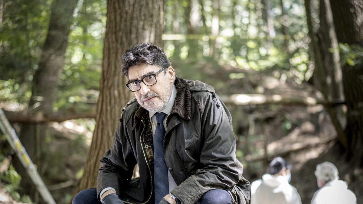 Alfred Molina as Detective Gamache crouching in a forest