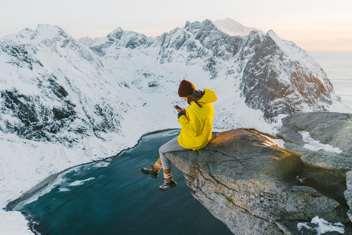 A woman in a yellow all-weather coat sits on a rock perched over a lake surrounded by snow-covered mountains.
