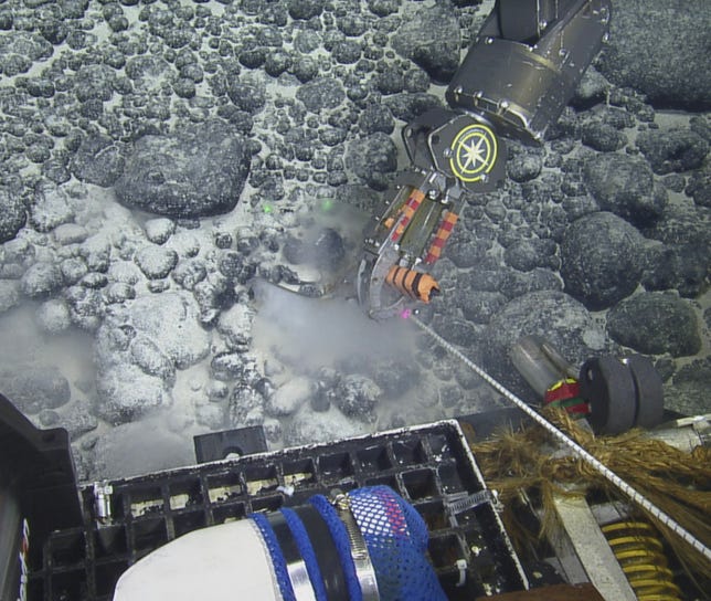 A gray, nodule-covered seamount in deep waters. A robotic vehicle scoops part of it up with an arm.