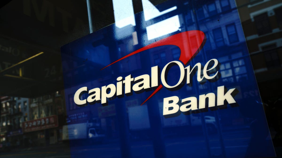 A red, white and blue sign featuring the Capital One name and a swoosh-like logo is displayed behind a window, set off by interesting reflections and shadows.